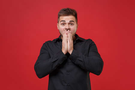 Shocked young bearded guy 20s in classic black shirt posing isolated on red wall background studio portrait. People sincere emotions lifestyle concept. Mock up copy space. Covering mouth with hands.の写真素材