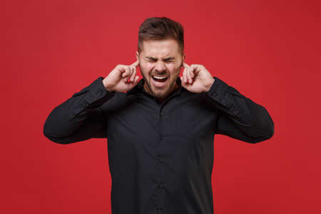 Crazy young bearded guy 20s in classic black shirt posing isolated on red background in studio. People lifestyle concept. Mock up copy space. Covering ears with fingers keeping eyes closed, screaming.の写真素材