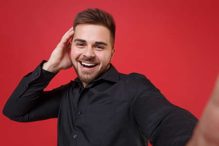 Close up of cheerful young bearded guy 20s in classic black shirt posing isolated on red background....