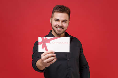 Smiling young bearded guy in classic black shirt posing isolated on bright red wall background studio portrait. People sincere emotions lifestyle concept. Mock up copy space. Hold gift certificate.の写真素材