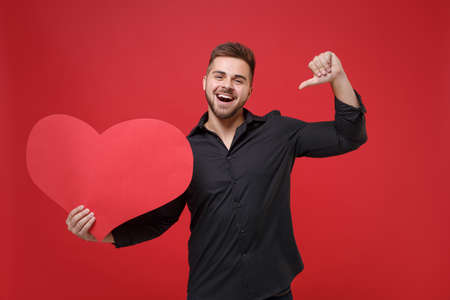 Cheerful young bearded guy in classic black shirt posing isolated on red wall background studio portrait. People lifestyle concept. Mock up copy space. Pointing thumb on big empty blank red heart.の写真素材