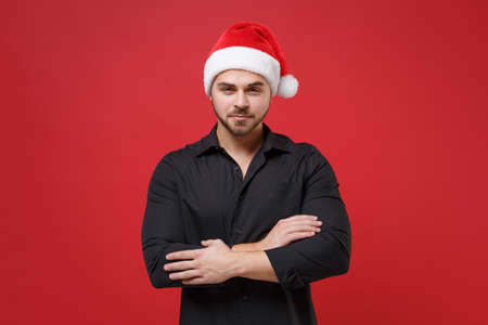 Handsome young bearded Santa man in classic black shirt, Christmas hat posing isolated on red background studio. Happy New Year celebration holiday concept. Mock up copy space. Holding hands crossed.の写真素材