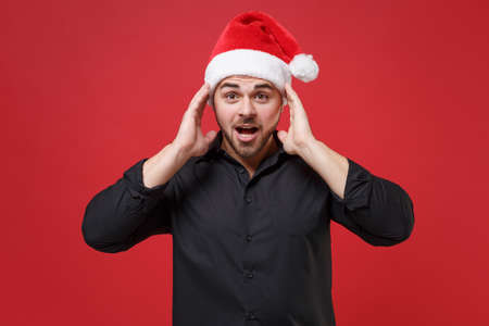 Shocked young bearded Santa man in classic black shirt Christmas hat isolated on red background. Happy New Year celebration holiday concept. Mock up copy space. Keeping mouth open, put hands on head.の写真素材