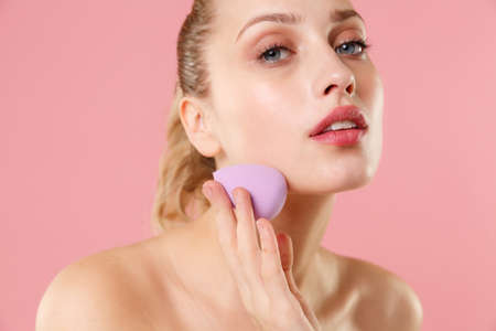 Close up blonde half naked woman 20s perfect skin blue eyes isolated on pastel pink wall background studio portrait. Skin care healthcare procedures concept Apply foundation nude makeup sponge on faceの写真素材