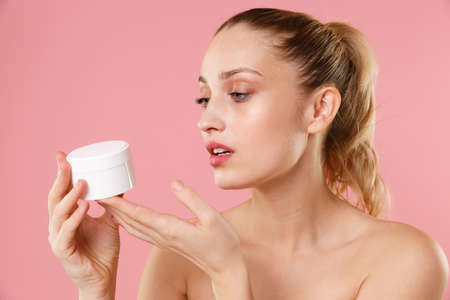 Close up blonde half naked woman 20s perfect skin nude makeup blue eyes isolated on pastel pink wall background. Skin care healthcare procedures concept Apply moisturizer, facial cream from container.の写真素材