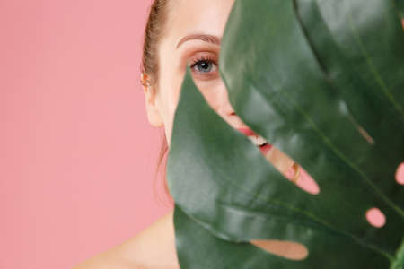 Close up blonde half naked woman 20s perfect skin nude make up blue eyes isolated on pastel pink background studio portrait. Skin care healthcare cosmetic procedures concept. Green tropical palm leaf.の写真素材