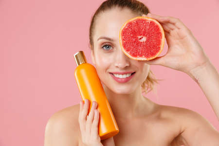 Close up half naked woman 20s perfect skin nude makeup blue eyes hold grapefruit bottle of shampoo isolated on pastel pink background studio portrait Skin care healthcare cosmetic procedures concept.の写真素材