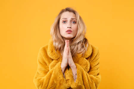 Pleading young blonde woman in yellow fur coat posing isolated on orange background in studio. People lifestyle concept. Mock up copy space. Holding hands folded in prayer, biting lips, looking aside.の写真素材