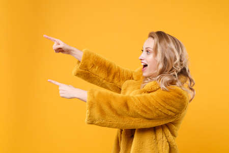 Side view of cheerful young blonde woman girl in yellow fur coat posing isolated on orange wall background studio portrait. People lifestyle concept. Mock up copy space. Pointing index fingers aside.の写真素材