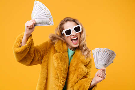 Happy young blonde woman girl in yellow fur coat, dark sunglasses isolated on orange background. People lifestyle concept. Mock up copy space. Holding fan of cash money in dollar banknotes, screaming.の写真素材
