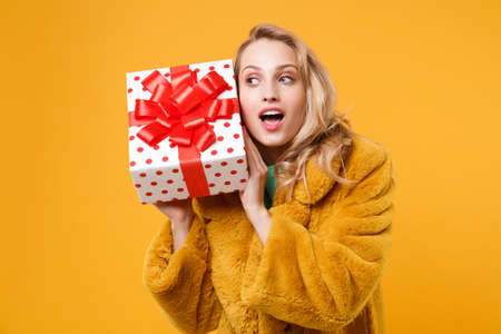Curious young woman girl in yellow fur coat isolated on orange background. St. Valentines Day, International Womens Day, birthday, holiday concept. Hold white red present box with gift ribbon bow.の写真素材