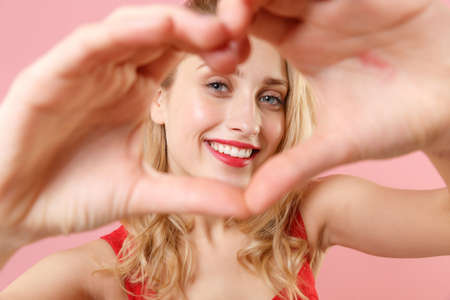 Smiling young blonde woman girl in red sexy clothes posing isolated on pastel pink background in studio. People lifestyle concept. Mock up copy space. Showing shape heart with hands, heart-shape sign.の写真素材