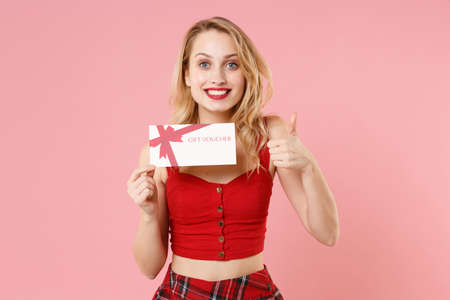 Smiling young blonde woman girl in red sexy clothes isolated on pastel pink background studio portrait. People emotions lifestyle concept. Mock up copy space. Hold gift certificate, showing thumb up.の写真素材