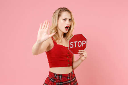 Dissatisfied young blonde woman girl in red sexy clothes isolated on pastel pink background. People lifestyle concept. Mock up copy space. Hold red sign with Stop title showing stop gesture with palm.の写真素材