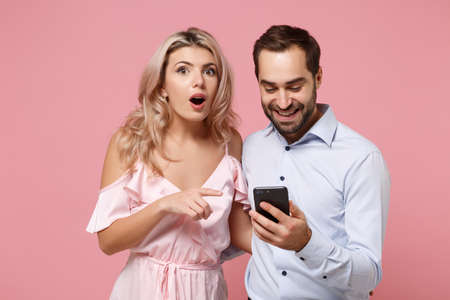 Amazed young couple two guy girl in party outfit celebrating isolated on pastel pink wall background. Valentines Day Womens Day birthday holiday party concept. Pointing index finger on mobile phone.の写真素材