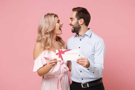Joyful young couple two guy girl in party outfit celebrating posing isolated on pastel pink background. People lifestyle Valentines Day, Womens Day birthday holiday concept. Hold gift certificate.の写真素材