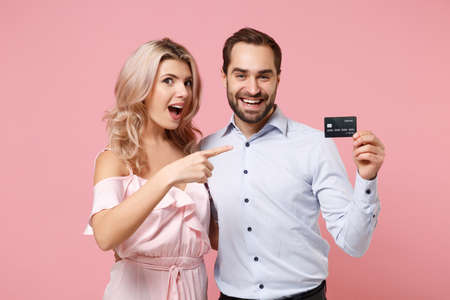 Excited young couple two guy girl in party outfit celebrating posing isolated on pastel pink background. Valentines Day, Womens Day birthday holiday concept. Point index finger on credit bank card.の写真素材