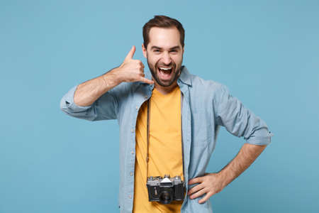 Traveler tourist man in yellow summer clothes with photo camera isolated on blue background. Male passenger traveling abroad on weekends. Air flight journey Doing phone gesture like says call me back.の写真素材