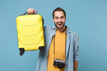 Cheerful traveler tourist man in yellow summer casual clothes with photo camera isolated on blue background. Male passenger traveling abroad on weekends. Air flight journey concept. Holding suitcase.の写真素材