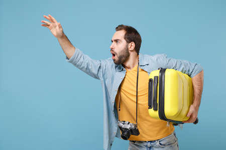 Traveler tourist man in yellow clothes with photo camera isolated on blue background. Male passenger travele abroad on weekends. Air flight journey. Hold suitcase waving hand for greeting, catch taxi.の写真素材