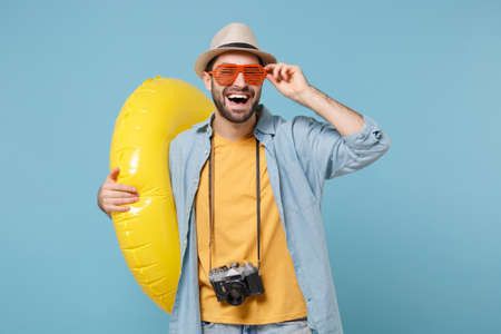 Laughing traveler tourist man in yellow clothes, eyeglasses with photo camera isolated on blue background. Passenger traveling abroad on weekends. Air flight journey concept. Hold inflatable ring.の写真素材