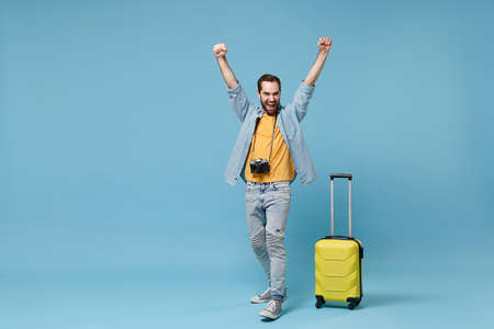 Happy traveler tourist man in yellow clothes with photo camera suitcase isolated on blue background. Male passenger traveling abroad on weekend. Air flight journey concept Clenching fists like winner.の写真素材
