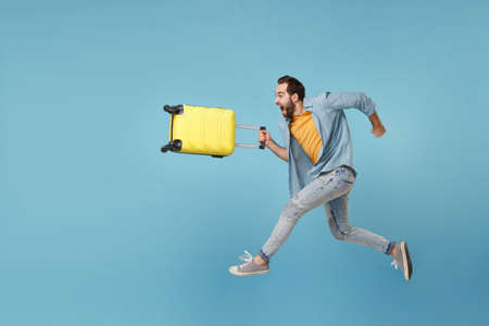 Side view of crazy traveler tourist man in yellow clothes isolated on blue background. Male passenger traveling abroad on weekend. Air flight journey concept. Jumping like running, hold suitcase.の写真素材