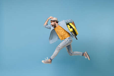 Side view of excited traveler tourist man in yellow clothes isolated on blue background. Male passenger traveling abroad on weekends. Air flight journey concept. Jumping like running, hold suitcase.の写真素材