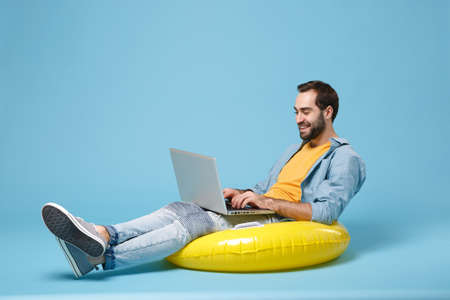 Smiling traveler tourist man in yellow clothes isolated on blue background. Passenger traveling abroad on weekend. Air flight journey concept. Sit in inflatable ring, work on laptop, booking hotel.の写真素材
