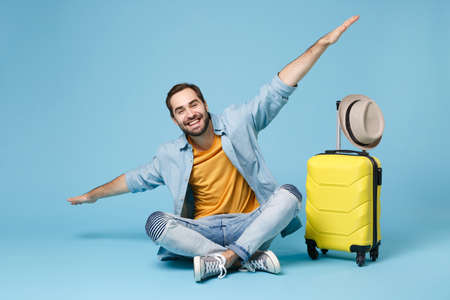 Smiling traveler tourist man in yellow clothes isolated on blue wall background. Male passenger traveling abroad on weekend. Air flight journey concept. Sit near suitcase, spreading hands like flying.の写真素材