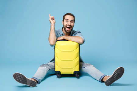 Excited traveler tourist man in summer clothes isolated on blue background. Male passenger traveling abroad on weekend. Air flight journey. Sit near suitcase, hold index finger up with great new idea.の写真素材