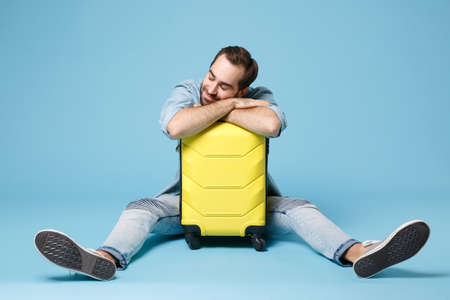 Smiling traveler tourist man in summer casual clothes isolated on blue wall background. Male passenger traveling abroad to travel on weekend. Air flight journey concept. Sitting sleeping on suitcase.の写真素材