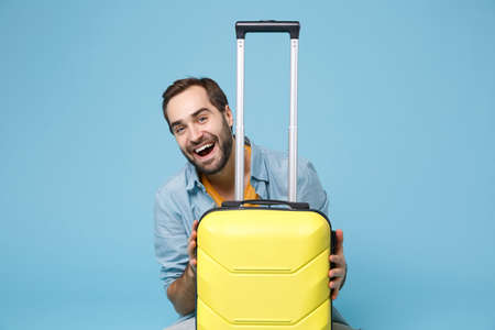 Cheerful traveler tourist man in summer yellow casual clothes isolated on blue wall background. Male passenger traveling abroad to travel on weekend. Air flight journey concept. Sitting near suitcase.の写真素材