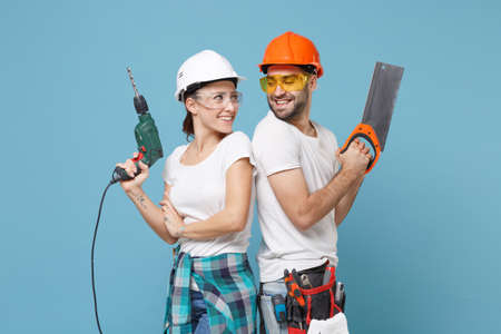 Smiling couple woman man in protective helmet hardhat hold electric drill saw isolated on blue background. Instruments accessories renovation apartment room. Repair home concept. Stand back to back.の写真素材