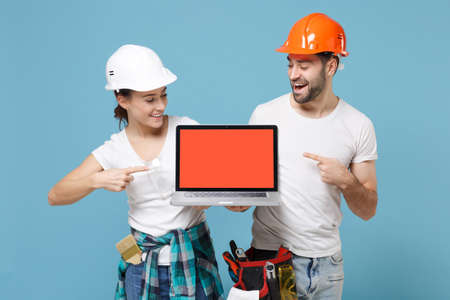 Cheerful couple woman man in hardhat point index fingers on laptop computer with blank empty screen isolated on blue background. Instruments accessories renovation apartment room. Repair home concept.の写真素材