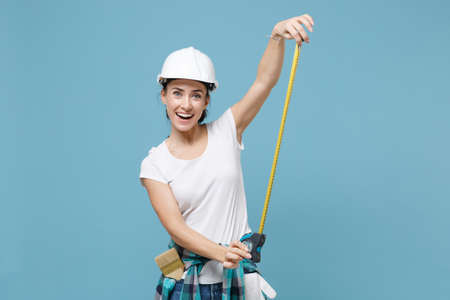 Excited young woman in casual clothes protective helmet hardhat hold measure tape isolated on blue background in studio. Instruments accessories for renovation apartment room. Repair home concept.の写真素材