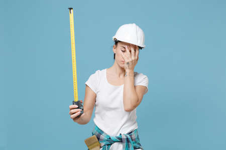 Exhausted young woman in protective helmet hardhat hold measure tape isolated on blue background studio. Instruments accessories for renovation apartment room. Repair home concept. Put hand on face.の写真素材