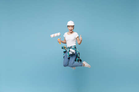 Excited young woman in protective helmet hardhat jump with paint roller isolated on blue wall...