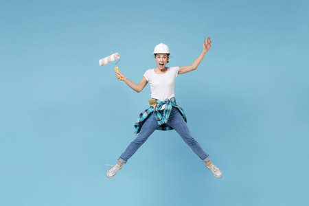 Excited young woman in protective helmet hardhat jump with paint roller isolated on blue background....