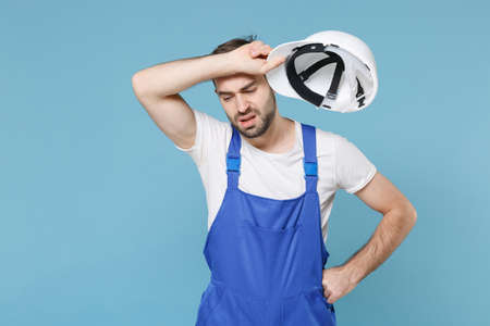 Exhausted young man in coveralls hold protective helmet hardhat isolated on pastel blue background...