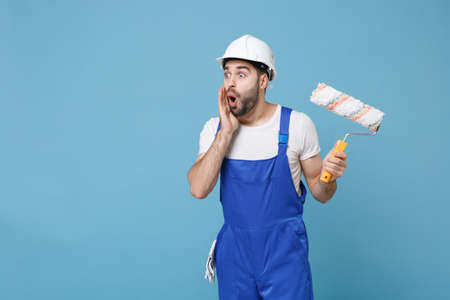 Shocked man in coveralls protective helmet hardhat hold paint roller isolated on blue background...