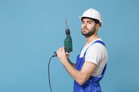 Side view serious young man in coveralls protective helmet hardhat hold electric drill isolated on pastel blue background. Instruments accessories for renovation apartment room. Repair home concept.の写真素材