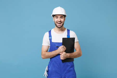 Funny young man in coveralls protective helmet hardhat hold clipboard with papers document isolated on blue wall background. Instruments accessories for renovation apartment room. Repair home concept.の写真素材