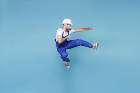 Funny young man in coveralls protective helmet hardhat isolated on blue background in studio....