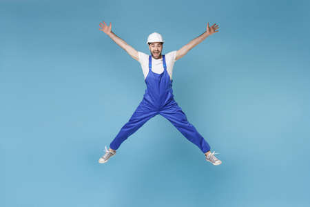 Excited young man in coveralls protective helmet hardhat isolated on blue background. Instruments accessories for renovation apartment room. Repair home concept. Jumping, spreading hands and legs.の写真素材
