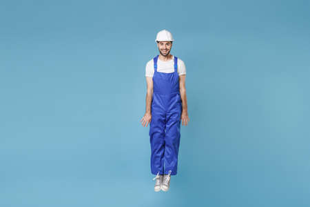 Smiling young man in coveralls protective helmet hardhat isolated on blue background studio portrait. Instruments accessories for renovation apartment room. Repair home concept. Jumping, having fun.の写真素材
