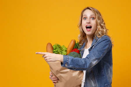 Amazed girl in denim clothes isolated on orange background. Delivery service from shop or restaurant concept Hold brown craft paper bag for takeaway mock up with food product point index finger aside.の写真素材