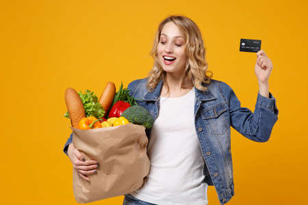 Funny young woman in denim clothes isolated on orange background. Delivery service from shop or restaurant concept. Hold brown craft paper bag for takeaway mock up with food product credit bank card.の写真素材