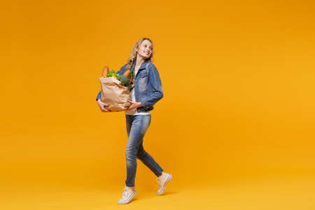 Smiling young woman in denim clothes isolated on orange background. Delivery service from shop or restaurant concept. Hold brown craft paper bag for takeaway mock up with food products looking aside.の写真素材