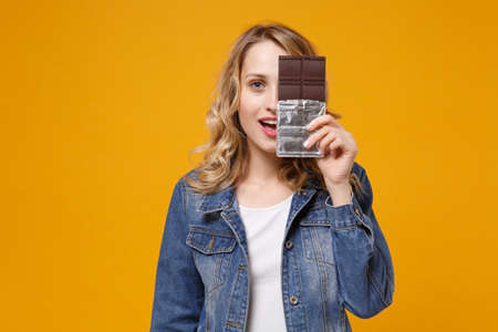 Funny young woman in denim clothes isolated on yellow orange background. Proper nutrition or sweets, dessert fast food, dieting morning concept. Mock up copy space. Covering face with chocolate bar.の写真素材